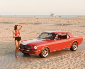 A woman in a red top and black shorts stands barefoot on a boardwalk at the beach, posing next to a classic red Ford Mustang coupe under warm sunset light, with the ocean and a lifeguard tower visible in the background